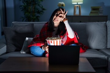 Sticker - Hispanic woman eating popcorn watching a movie on the sofa doing ok gesture with hand smiling, eye looking through fingers with happy face.