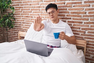 Wall Mural - Young asian man drinking coffee sitting on the bed with open hand doing stop sign with serious and confident expression, defense gesture