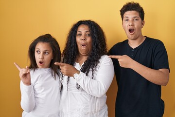 Poster - Family of mother, daughter and son standing over yellow background surprised pointing with finger to the side, open mouth amazed expression.