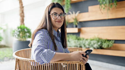 Wall Mural - Young beautiful hispanic woman smiling using smartphone sitting on a chair at the office garden