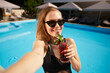 © artiemedvedev - Travel blogger woman in bikini taking selfie photo with cocktail near swimming pool. Pov of lifestyle vlogger selfie from vacation on luxury resort. Female in swimwear drinks beverage on tropical spa.