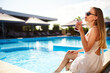 © artiemedvedev - Blonde woman drinks refreshing cocktail sunbathing and sitting near swimming pool at tropical spa. Female in sunglasses enjoys a drink on poolside. Girl chilling in tropical resort on vacation.