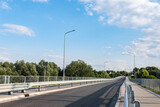 Fototapeta Miasto - Oświetlenie drogi w obszarze wiejskim, na pierwszym planie barierki ochronne, w tle drzewa i błękitne niemalże bezchmurne niebo