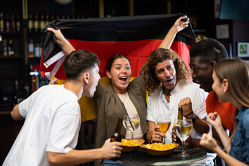 Wall Mural - Happy multiracial sport fans holding up in a flag of Germany and watching football competitions with beer in the pub