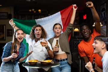 Wall Mural - Happy multiracial friends holding the flag of Italy, rejoicing winning football game and drinking beer in the bar