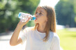 © Irina Mikhailichenko - Portrait of beautiful teenager pretty girl, young thirsty woman is drinking pure fresh water from bottle at warm sunny summer day outdoors in a green park.
