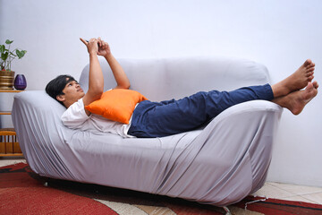 Full length view of Asian man with smartphone laying on the sofa at home. Online chatting on phone, scrolling news feed, spends time in social networks