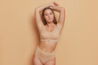 Graceful young woman wearing beige bra and panties stands at light brown background, hands over her head, natural beauty concept, copy space