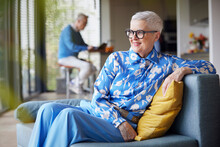 Relaxed Senior Woman Sitting On Couch At Home With Man In Background