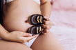 Pregnant woman strokes hugging the belly tummy abdomen enjoying pregnancy sitting on bed dressed underwear hold child shoes. Future family, baby infant expecting child inside
