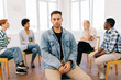 © dikushin - Portrait of sad Caucasian man looking at camera with serious expression while group of multicultural and different ages people sitting in circle, talking in background during therapy session.