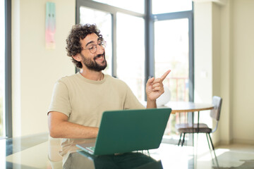 Sticker - young adult bearded man with a laptop smiling cheerfully, feeling happy and pointing to the side and upwards, showing object in copy space