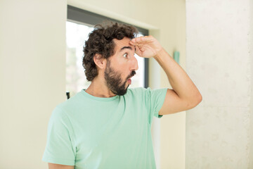 Sticker - young crazy bearded man looking bewildered and astonished, with hand over forehead looking far away, watching or searching