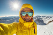 Frau in gelber Winterjacke und Skibrille macht ein Selfie mit schneebedeckten Bergen im Hintergrund im Sonnenschein