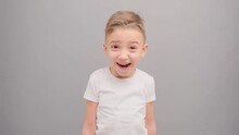 A Little Boy In A White T-shirt Looks Thoughtfully Into The Camera And Then Smiles Happily. Stop Motion Filmed In The Studio On A Gray Background