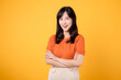 © Jirawatfoto - Step into confident grace with a young Asian woman in her 30s, wearing an orange shirt, displaying crossed arm sign gesture on yellow background.