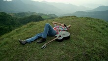 Male Musician With Acoustic Guitar Lays Down At Green Mountain Hills Landscape Watching The Sky, Wearing Vintage Colorful Clothes