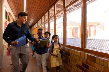 Wall Mural - Diverse male teacher and children walking together in corridor at elementary school, copy space