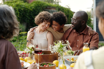 Happy boy embracing his intercultural parents sitting by table with homemade food while enjoying outdoor family dinner on weekend