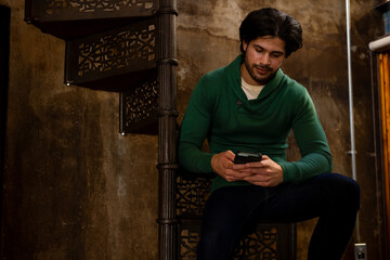 Indian man sitting on stairs and using smartphone at home, copy space