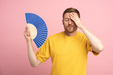 Too Hot. Sweaty Tired Middle Aged Man Touching Forehead Using Paper Fan Suffer From Heat, Feels Sluggish. Displeased Guy Cooling In Hot Summer Weather, Isolated On Studio Pink Background. Overheating