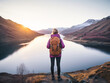 A young blonde female hiker wearing a purple down jacket and orange backpack looks out onto a lake at dawn. Her back is to the camera. Snowcapped mountains in the distance.