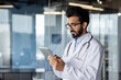 © Tetiana - A young Indian doctor works at a tablet, stands in a white coat with a stethoscope in the hospital lobby