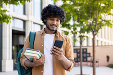 Young Indian Male Student Using Mobile Phone, Standing Outside On Street, Holding Books In Hand, Reading Message, Looking For Campus, Waiting For Meeting