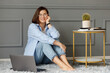 © JJ Studio - Pensive businesswoman, wearing glasses on her eyes, is wearing white t-shirt, blue shirt and jeans, sits holding her hand on her chin on a gray carpet on the floor in front of laptop