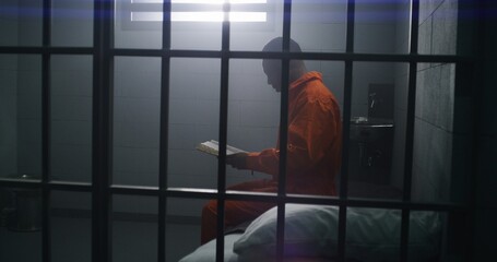 Wall Mural - African American prisoner in orange uniform sits on the bed, reads Bible in prison cell. Male criminal serves imprisonment term for crime in jail or detention center. Faith in God. View through bars.