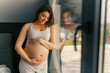 © DusanJelicic - a pregnant woman with long brown hair stands leaning against the window with her hands gently on her stomach and looking down