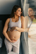 © DusanJelicic - a pregnant woman with long brown hair stands leaning against the window with her hands gently on her stomach and looking out the window