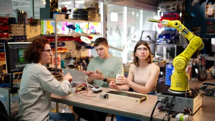 Young happy engineers programming an yellow robotic arm in the workshop to grab cardboard water glass, computer programming training for coffee preparation, celebrating success