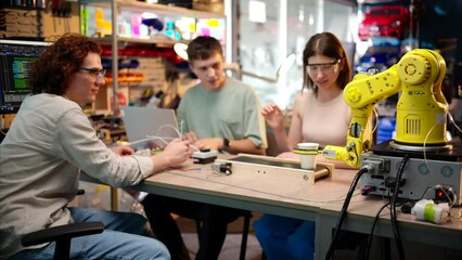Young happy engineers programming an yellow robotic arm in the workshop to grab cardboard water glass, computer programming training for coffee preparation, celebrating success
