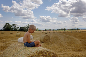 Wall Mural - Beautiful blond child, boy, lying on a haystack in the field. Amazing landscape, rural scene with clouds, tree and empty road summertime, fields of haystack next to the road