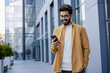 © Liubomir - Young successful businessman walking in the city outside office building, hispanic smiling happy with achievement results, holding phone, using online application outdoors.