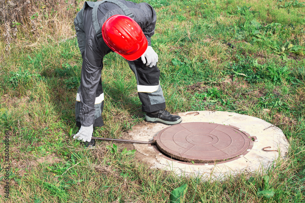 Manhole Root Control A worker in overalls and a helmet opens a manhole with a crowbar ...