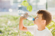 © johnalexandr - A beautiful child sitting on the grass drinks water from a bottle in the summer at sunset. Boy quenches his thirst on a hot day