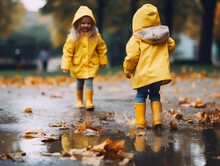 Two Little Girls In Yellow Jackets And Rubber Boots. Children Jump And Play In The Rain Puddle. Yellow, Colorful Leaves. Autumn Trees.