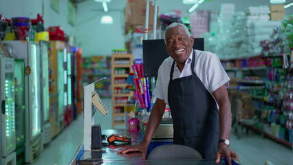 One happy black senior employee standing at supermarket cashier smiling at camera wearing apron uniform. Charismatic friendly African American staff of Grocery store