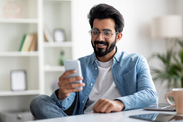 Wall Mural - Great App. Smiling Indian Man Using Smartphone At Home Office