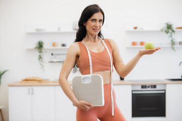 Wall Mural - Slender caucasian woman with measuring tape and scales holding apple on palm while standing in kitchen. Effective health coach in sportswear assisting in weight management via healthy nutrition.