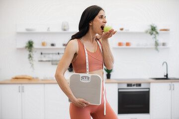 Healthy female with measuring tape biting ripe fruit after checking weight on scales in home interior. Pretty woman in sports clothes staying in good shape due to physical exercises and balanced meal.