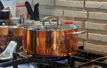 Copper Saucepan With Lid With Dish Is Cooked Over Fire On Gas Stove. Steam Rises Above The Pot