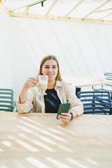 Wall Mural - Happy woman sitting at outdoor cafe table and talking on phone with cup of coffee, smiling woman enjoying telecommuting in cafe or studying online