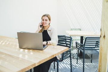 Wall Mural - Young happy woman sitting at outdoor cafe table and talking on phone with cup of coffee, smiling woman enjoying telecommuting in cafe or studying online on laptop