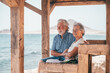 © luciano - Lovely senior family couple sitting close to the beach looking at horizon over sea, two elderly people enjoying vacation or retirement together