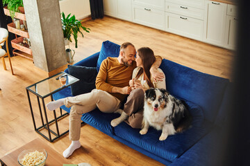 Wall Mural - Smiling man hugging girlfriend with coffee while sitting near border collie on couch at home