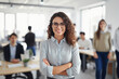 Smiling hipster woman in modern office, arms crossed, colleagues in background.