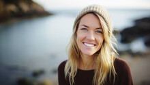 jeune femme blonde de type nordique souriante avec un bonnet en laine au bord de la mer ou d'un lac en arrière plan